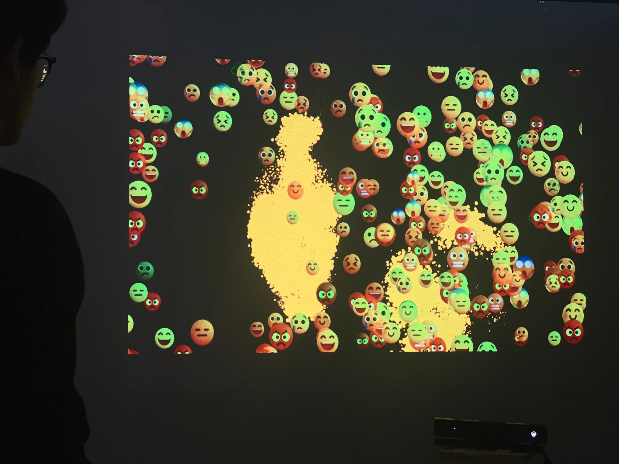 The installation seen from the back of the room. A visitor stands in silhouette at the left edge, the projection covers the wall in front of them with emoji around a yellow particle figure, and the Kinect sits on a low stand below the image.