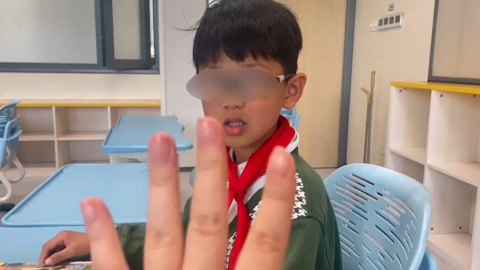 A seated pupil in a red scarf, face blurred, with another person’s hand raised in front of the camera.