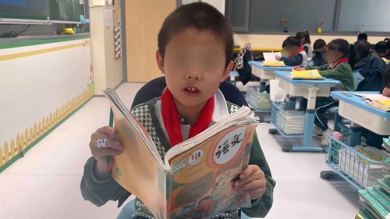 A pupil reading a Chinese language textbook aloud, face blurred, with classmates working at their desks behind.