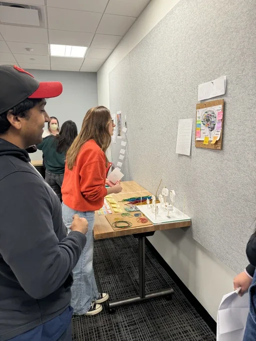 A testing session in a corridor. Two people approach a table holding the prototype brain, bundles of coloured rubber bands and a spread of cards; a printed board with sticky notes hangs on the wall behind.