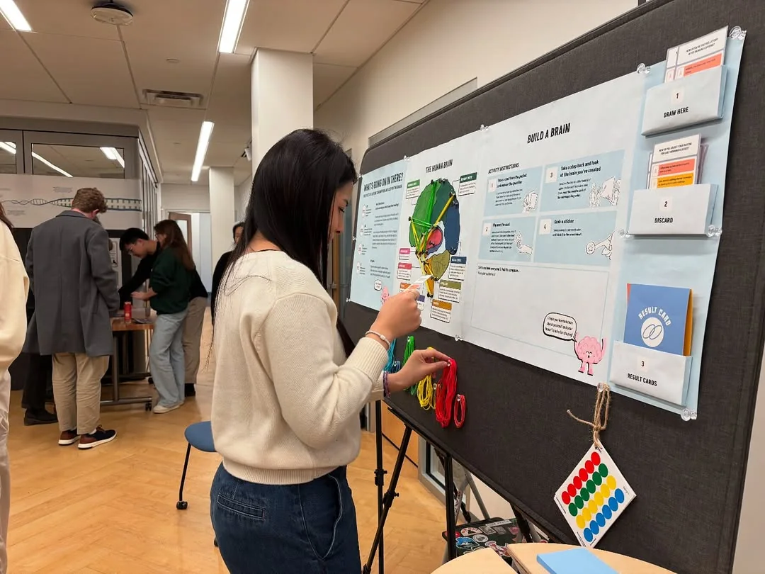 A visitor standing at the board mid-activity, holding a red rubber band and reaching towards the pins, with the card holders and the sticker key beside her.